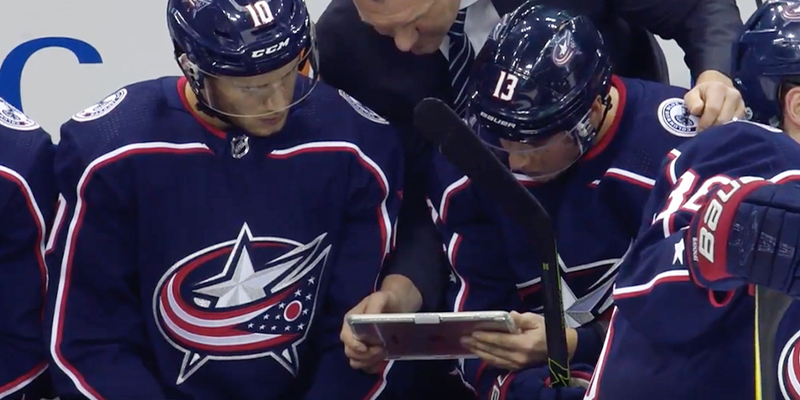 Two players from the Columbus Blue Jackets look at an iPad while sitting on the bench.
